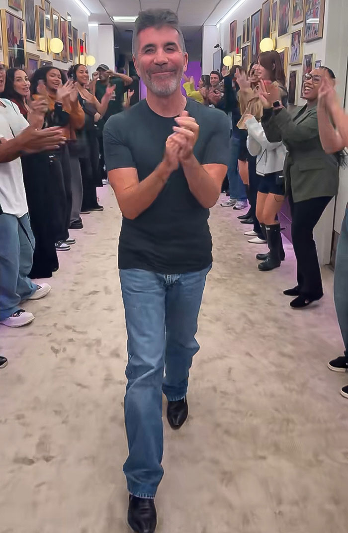 Simon Cowell walking and clapping in a hallway, surrounded by people applauding after bike accident survival.