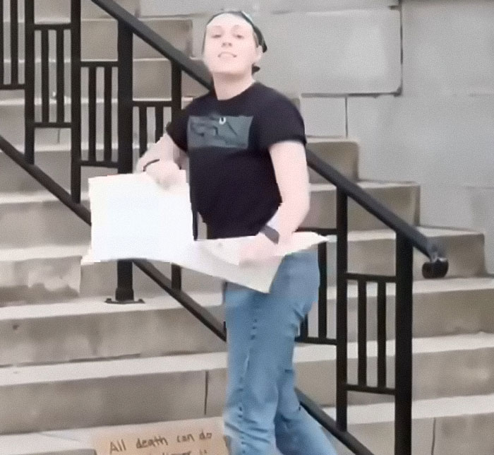Young woman standing on stairs holding cardboard sign, associated with sisters who destroyed Charlie Kirk memorial.