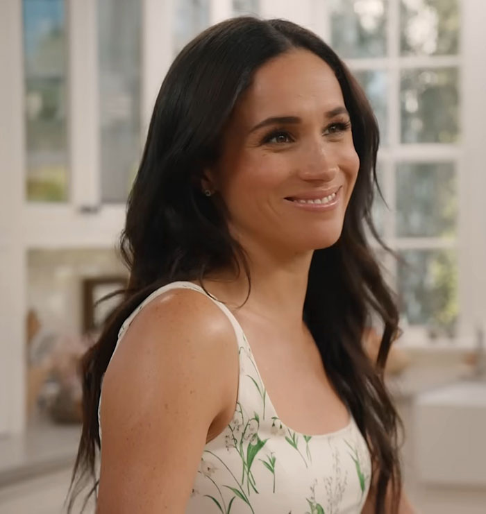 Meghan Markle smiling indoors wearing a white floral dress with long dark hair, looking content and relaxed.