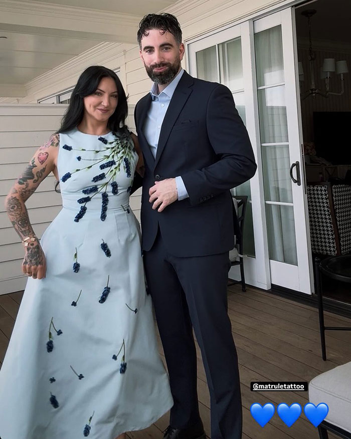 Woman in a light blue floral dress and tattoo sleeves, standing next to a man in a dark suit at a wedding event.