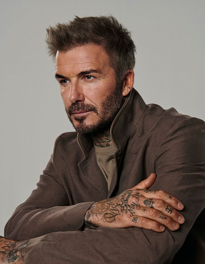 David Beckham with tattoos on his body, posing thoughtfully in a brown jacket, highlighting signs seen by a doctor.