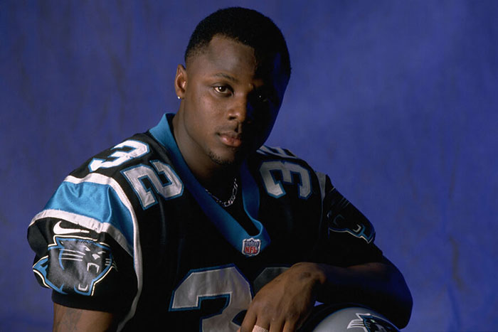 Male celebrity in a Carolina Panthers NFL jersey posing against a blue backdrop related to slain male celebrities by women.