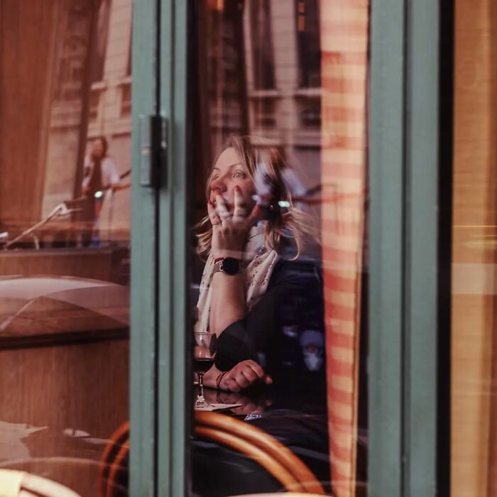 Woman sitting indoors at a café, seen through glass with reflections, captured in stunning street photos by Greek photographer.