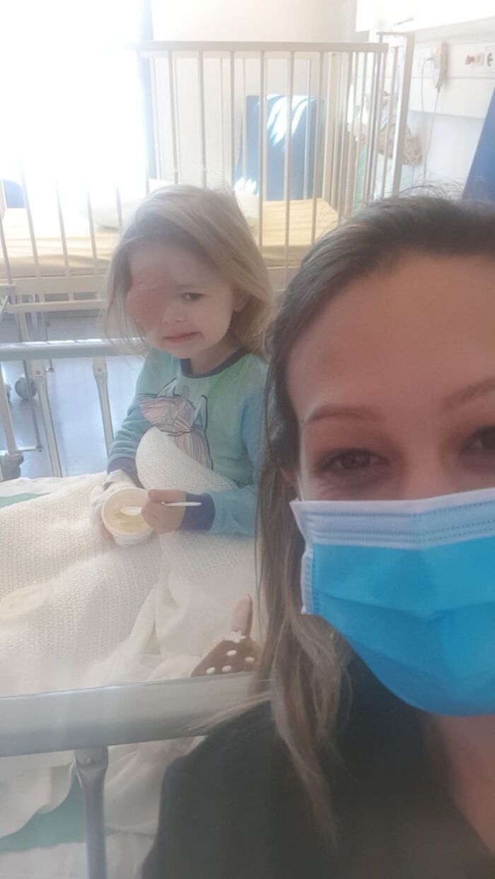 Young child wearing eye patch eating in hospital bed while woman in blue mask takes a selfie, wholesome posts to distract the world.