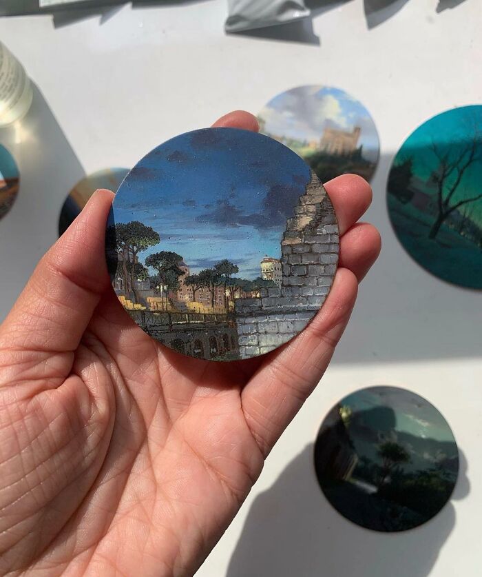 Hand holding a detailed round painting by Dina Brodsky depicting trees and architecture under a twilight sky.