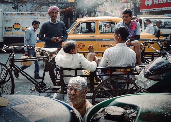 Street photo showing a busy urban scene with men, a yellow taxi, bicycles, and a man covered in soap in a crowded street.
