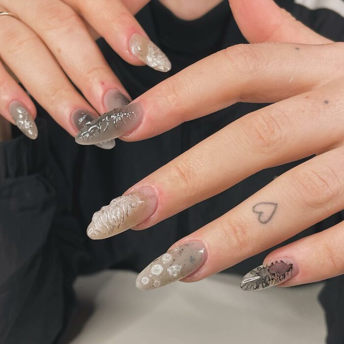 Long almond-shaped nail designs with textured gray and beige patterns on hands with a small heart tattoo, showcasing unique nail designs.