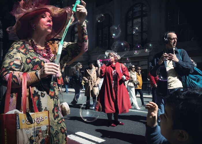 Woman in a colorful dress and hat blowing bubbles on a busy city street among pedestrians in a striking street photo.