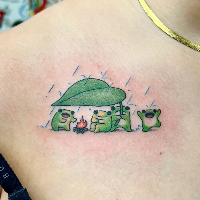 Small adorable frog tattoo by Korean artist Buoy showing frogs sheltering under a leaf and sitting by a fire in rain.