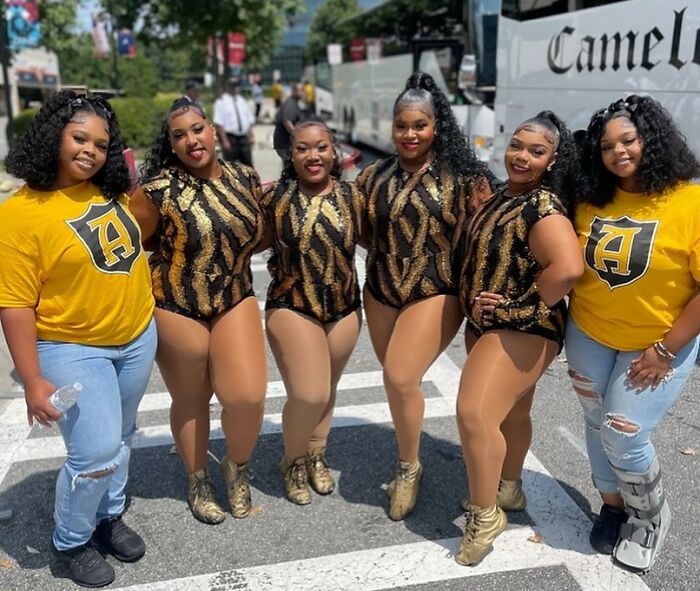 Plus-size college band dancers pose together outdoors in matching gold and black performance outfits at halftime event.