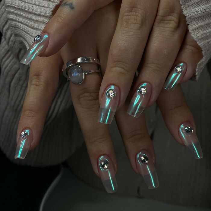 Long iridescent nail designs with metallic rose embellishments on polished clear acrylic nails.