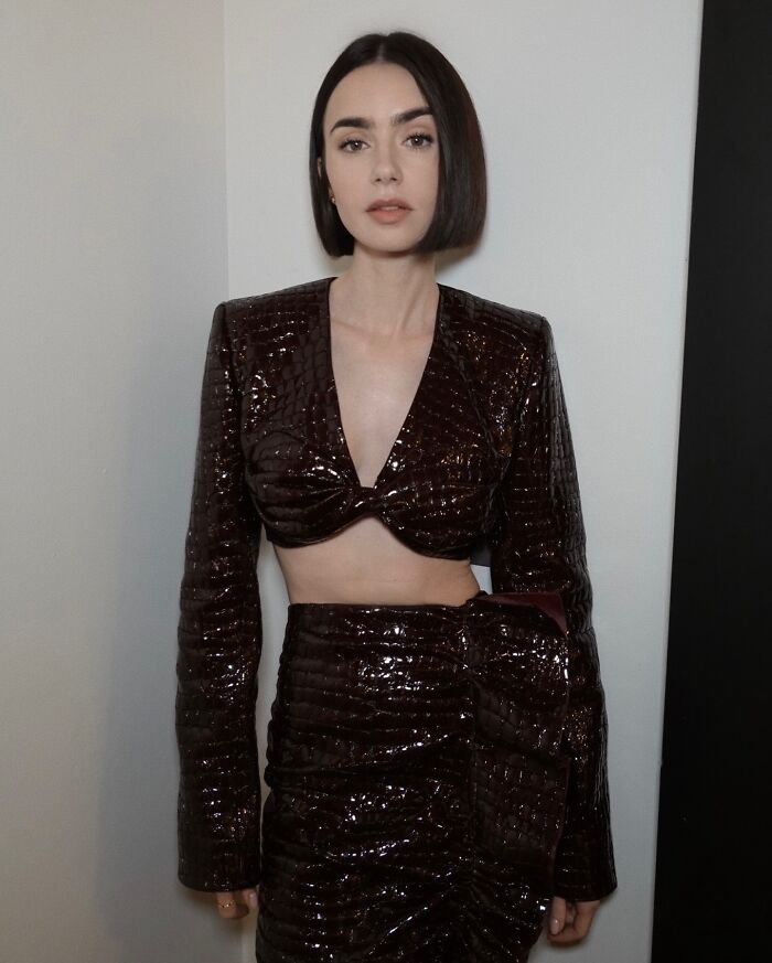 Lily Collins posing in a shiny dark cropped top and skirt, highlighting her extreme thinness and slender figure.