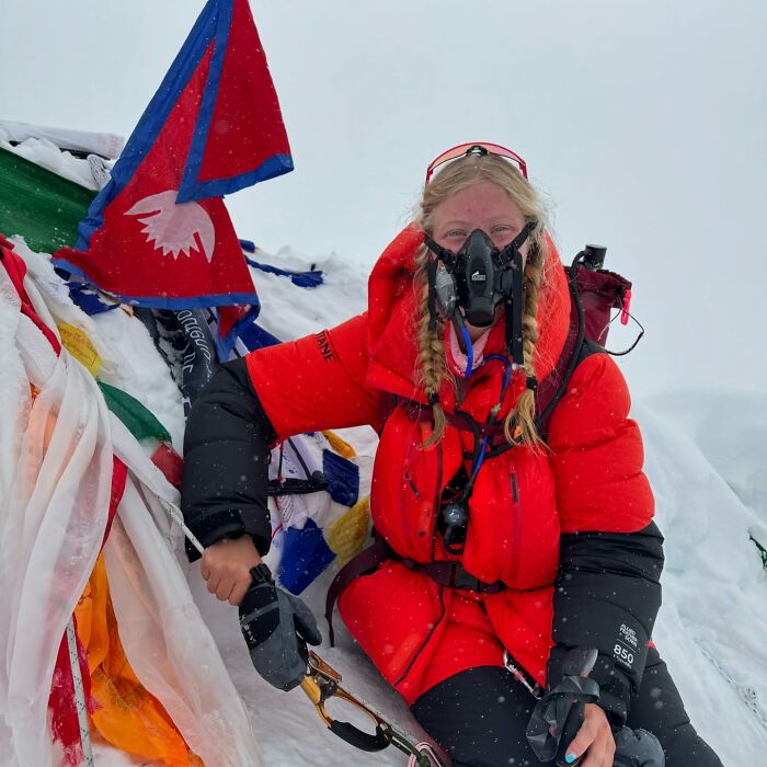 Teen climber in red jacket wearing oxygen mask on snowy Everest summit amid flags, highlighting Everest climb risk debate. Teen climber in red jacket wearing oxygen mask on snowy Everest summit amid flags, highlighting Everest climb risk debate.