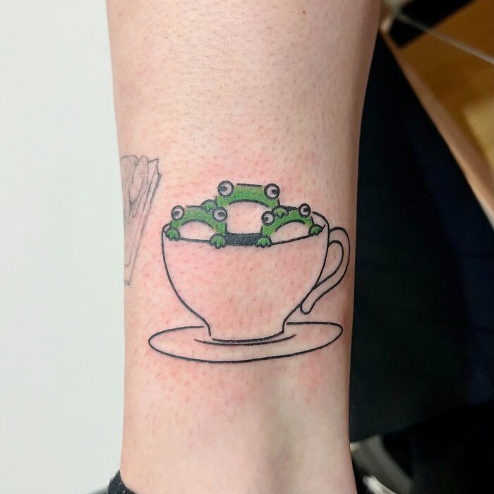 Tattoo of adorable frogs sitting inside a teacup, showcasing cute animal tattoo art by Korean artist Buoy.
