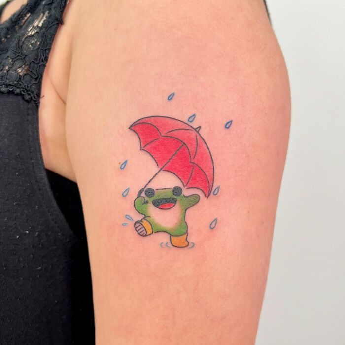Small adorable frog tattoo holding a red umbrella with raindrops, colorful animal tattoo by Korean artist Buoy on upper arm.