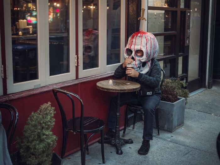 Person wearing a blood-streaked skull mask sitting at a small table outdoors in a striking street photo scene.