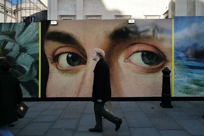 Person walking past street photo mural with giant eyes, capturing the funny side of life in an urban setting.