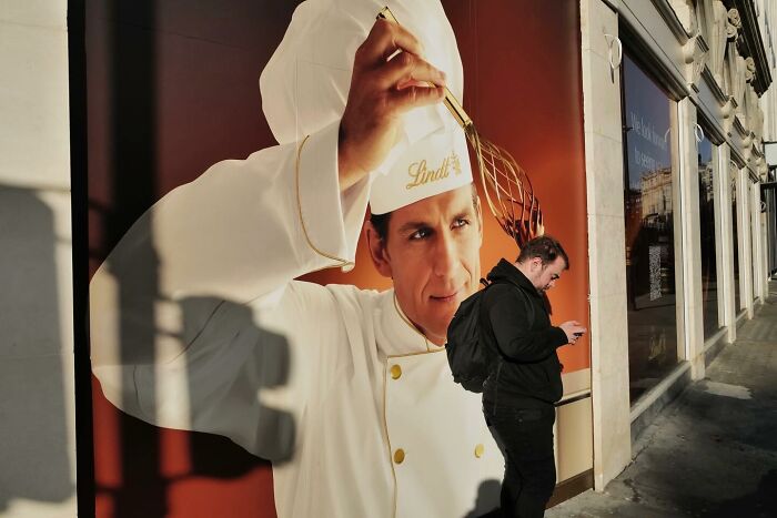 Man standing by a large humorous street photo of a chef whisking, capturing the funny side of life in urban scenes.