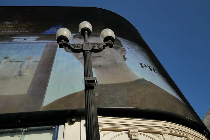 Black lamp post stands in front of a large curved billboard under a clear blue sky in a street photo capturing the funny side of life.
