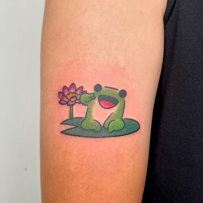 Cute frog tattoo with a pink lotus flower, showcasing adorable animal tattoo style by Korean artist Buoy on upper arm.