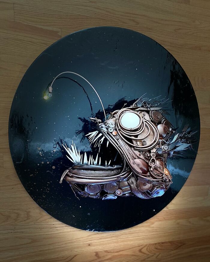 Stunning animal sculpture made from trash depicting an anglerfish with intricate mechanical details on a black circular background.