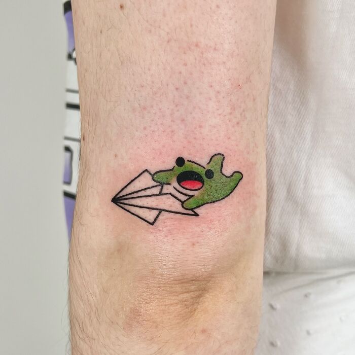 Green adorable frog tattoo on forearm holding a paper airplane, created by Korean artist Buoy in a colorful style.