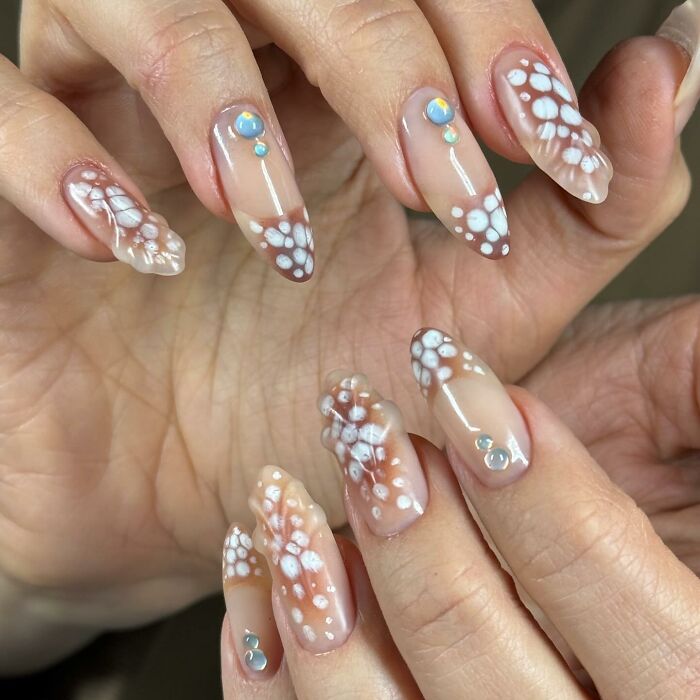 Nail designs featuring translucent brown tips with white floral patterns and small blue gemstone accents on long nails.