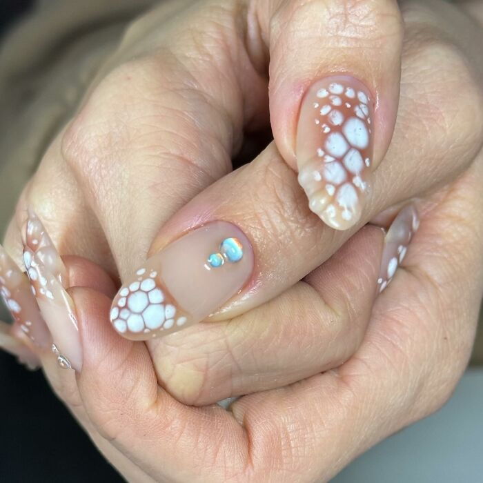 Nail designs featuring translucent brown tips with white floral patterns and small blue gemstone accents on long nails.