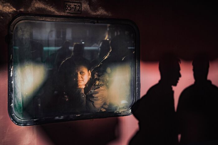 Woman in a jacket looking through a train window with silhouettes of people outside in striking street photo.