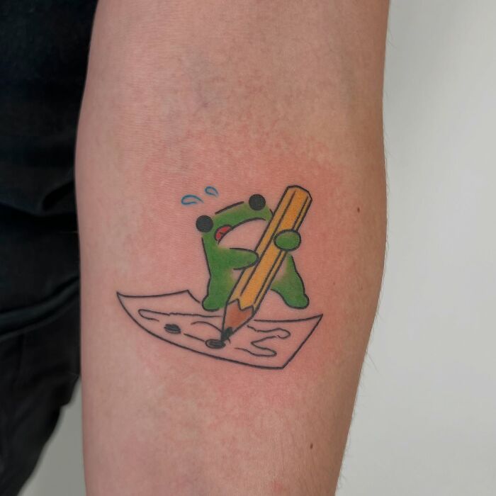 Adorable frog tattoo holding a pencil, creating a drawing, colorful animal tattoo by Korean artist Buoy on forearm.