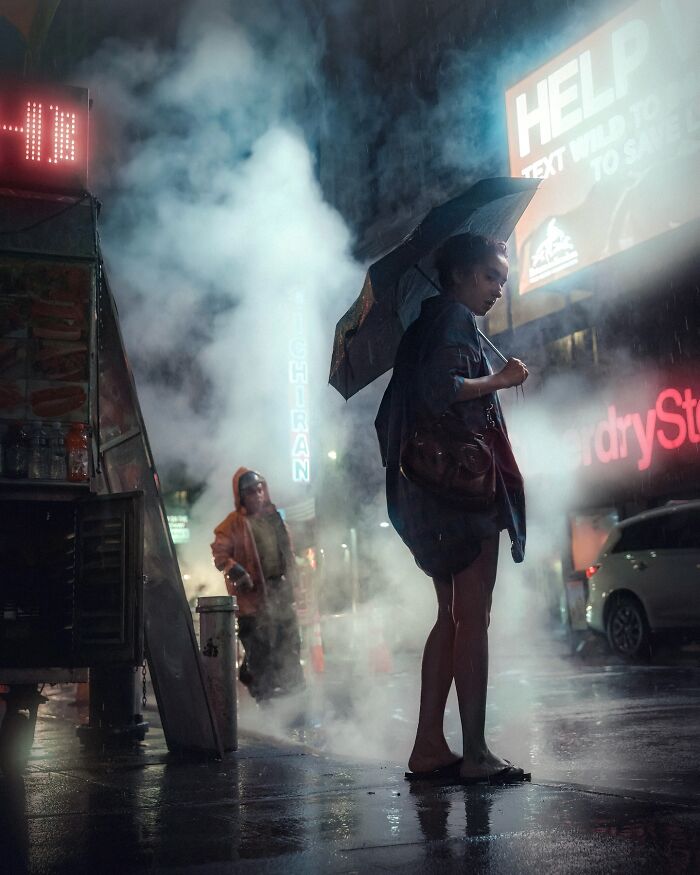Woman standing on rainy street at night with umbrella and illuminated signs in background, striking street photo like movie still.