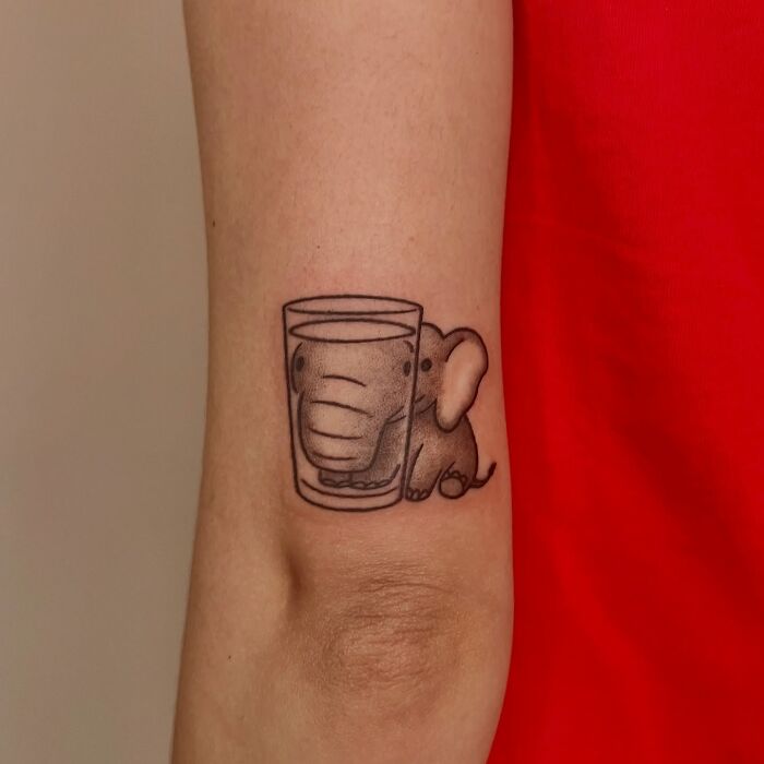 Small elephant tattoo behind a glass of water on forearm, an adorable animal tattoo by Korean artist Buoy.