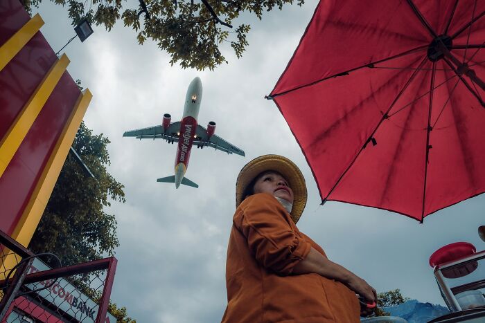 Person wearing a hat beneath a red umbrella with a plane flying overhead in a striking street photo scene.