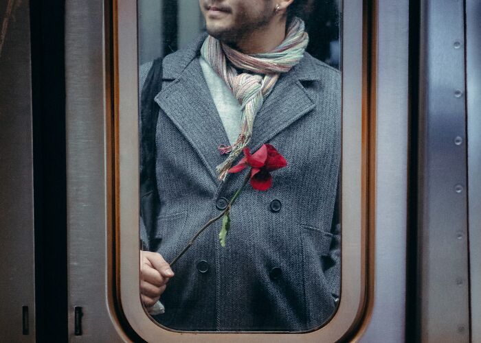 Man in gray coat and scarf holding a red flower inside a subway train in a striking street photo scene.