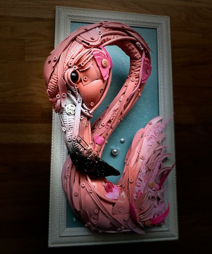Pink flamingo animal sculpture made from repurposed trash items, displayed in an ornate white frame against a wooden background.