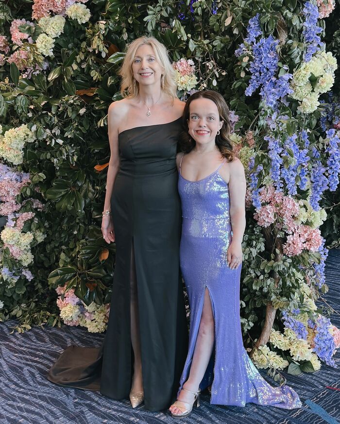 31YO woman with dwarfism in a sparkling purple gown posing with a taller woman in a black dress against a floral backdrop