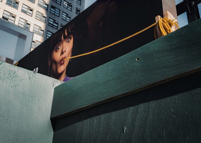 Street photo of a distorted face on a billboard with a yellow cable plugged into the mouth, captured in an urban setting.
