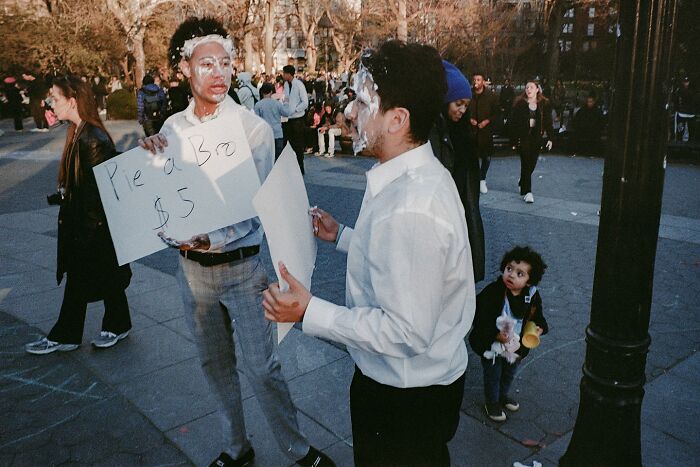 Two men with faces covered in pie cream holding signs in a busy urban street scene, striking street photos style.