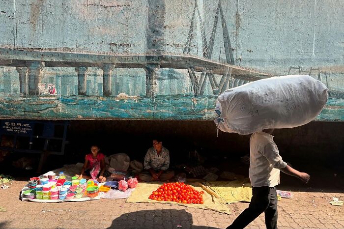 Street photo showing a man carrying a large sack past vendors selling colorful bowls and tomatoes under a painted bridge.
