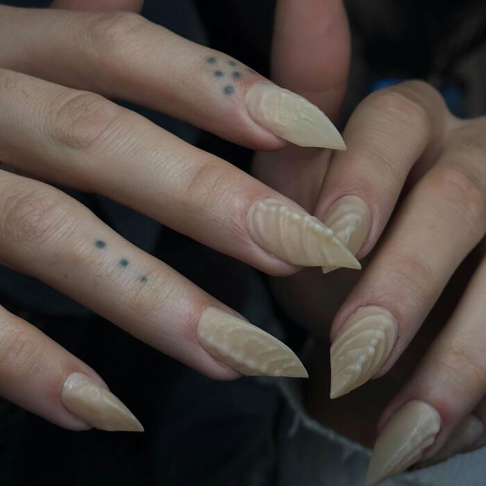 Long n**e almond-shaped nail designs with textured patterns on fingers featuring small dot tattoos, showcasing bold nail designs.