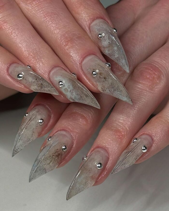 Long pointed nail designs with marble-like texture and silver studs, showcasing creative and unique nail art styles.