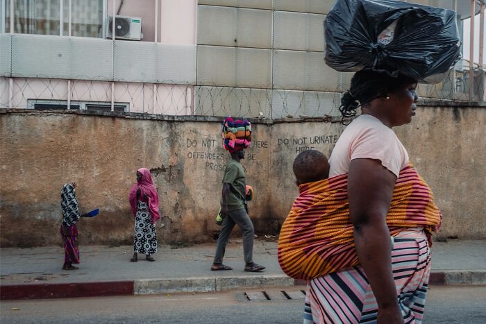 Street photo of a woman carrying a child and a bag on her head walking past pedestrians on an urban sidewalk.