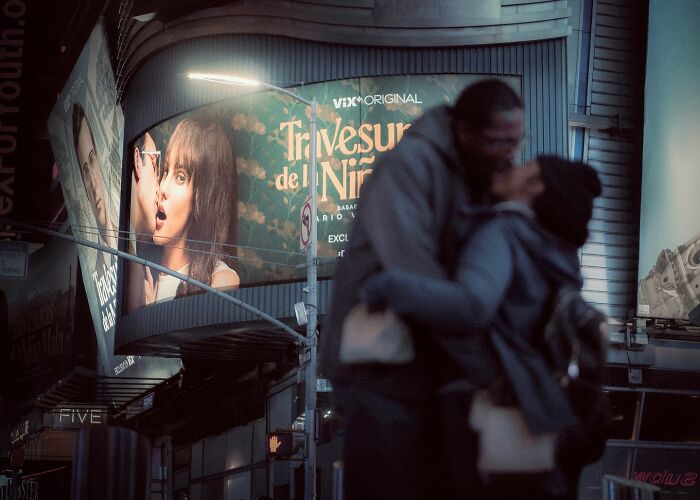 Couple embracing on a city street at night with illuminated movie-like street photos in the background.