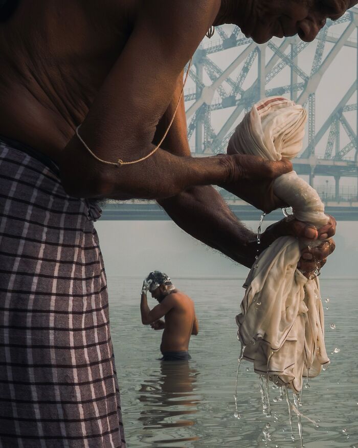 Man washing clothes in river near bridge with another man bathing, striking street photos with cinematic feel
