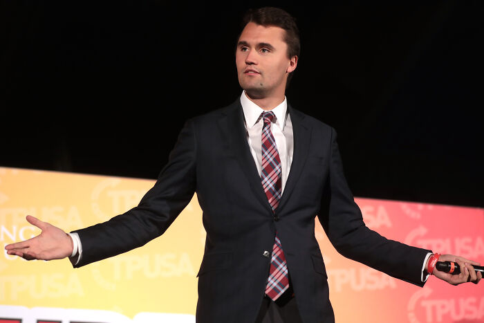 Charlie Kirk speaking at an event, wearing a suit and tie with arms outstretched against a colorful backdrop.