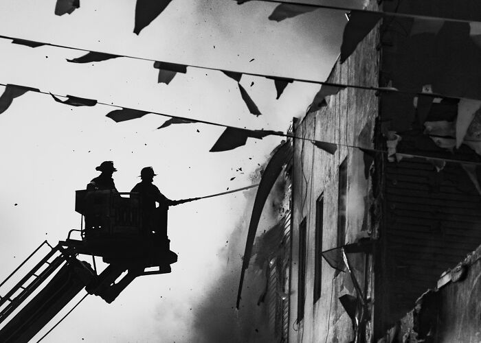 Two firefighters on a lift spraying water on a burning building in a striking street photo that feels like a movie still.