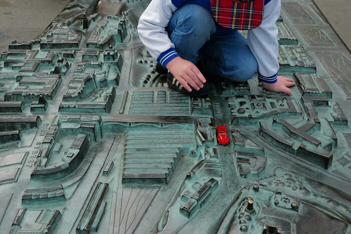 Child interacting with a detailed bronze street map sculpture, capturing a funny side of life in a street photo.