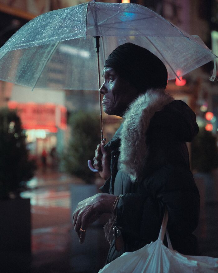 Elderly person holding a transparent umbrella on a rainy city street, captured in a striking street photo with cinematic lighting.