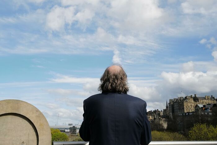 Man with thinning hair stands on a balcony overlooking city buildings and cloudy sky in a funny street photo.