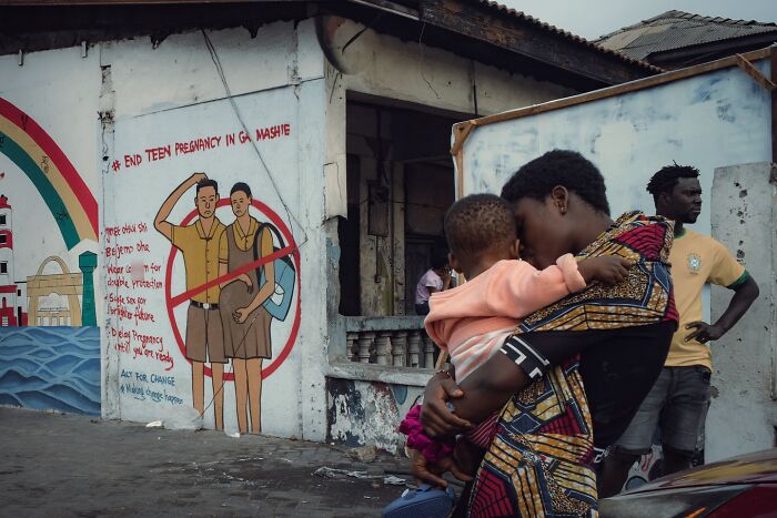 Street photo of a woman holding a child near a mural against teen pregnancy in an urban setting, striking street photos style.